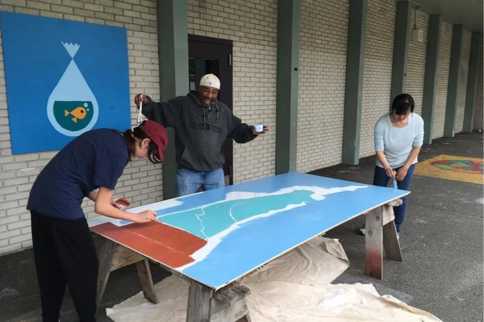 Volunteers painting a large mural panel outside the school